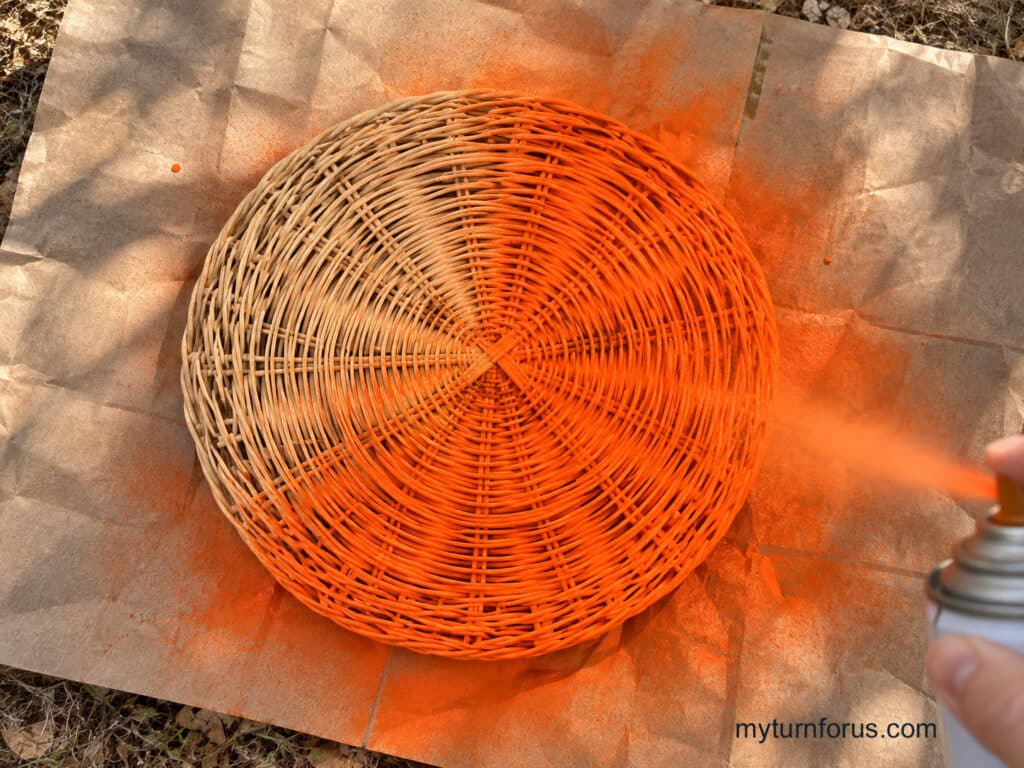 Overhead view of a woven straw paper plate holder being spray painted orange on a protected outdoor surface for a pumpkin witch Halloween decoration.