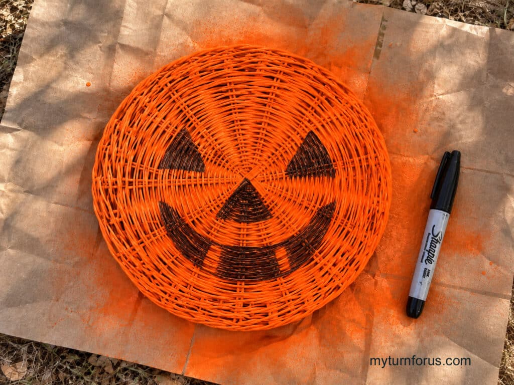 Overhead view of an orange painted woven straw paper plate holder with a jack-o’-lantern face drawn on it and a black marker beside it on a paint-covered outdoor work surface.