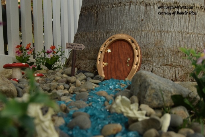 Horseshoe Fairy Door for the most magical fairy garden! https://www.myturnforus.com/2016/03/horseshoe-fairy-door.html Golden horseshoe fairy door in a small garden scene with a blue glass path, pebbles, flowers, mushrooms, and a “Fairies Welcome” sign.