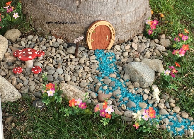 Horseshoe Fairy Door for the most magical fairy garden! Golden horseshoe fairy door displayed at the base of a tree with a blue glass path, river rocks, red mushrooms, and colorful flowers.