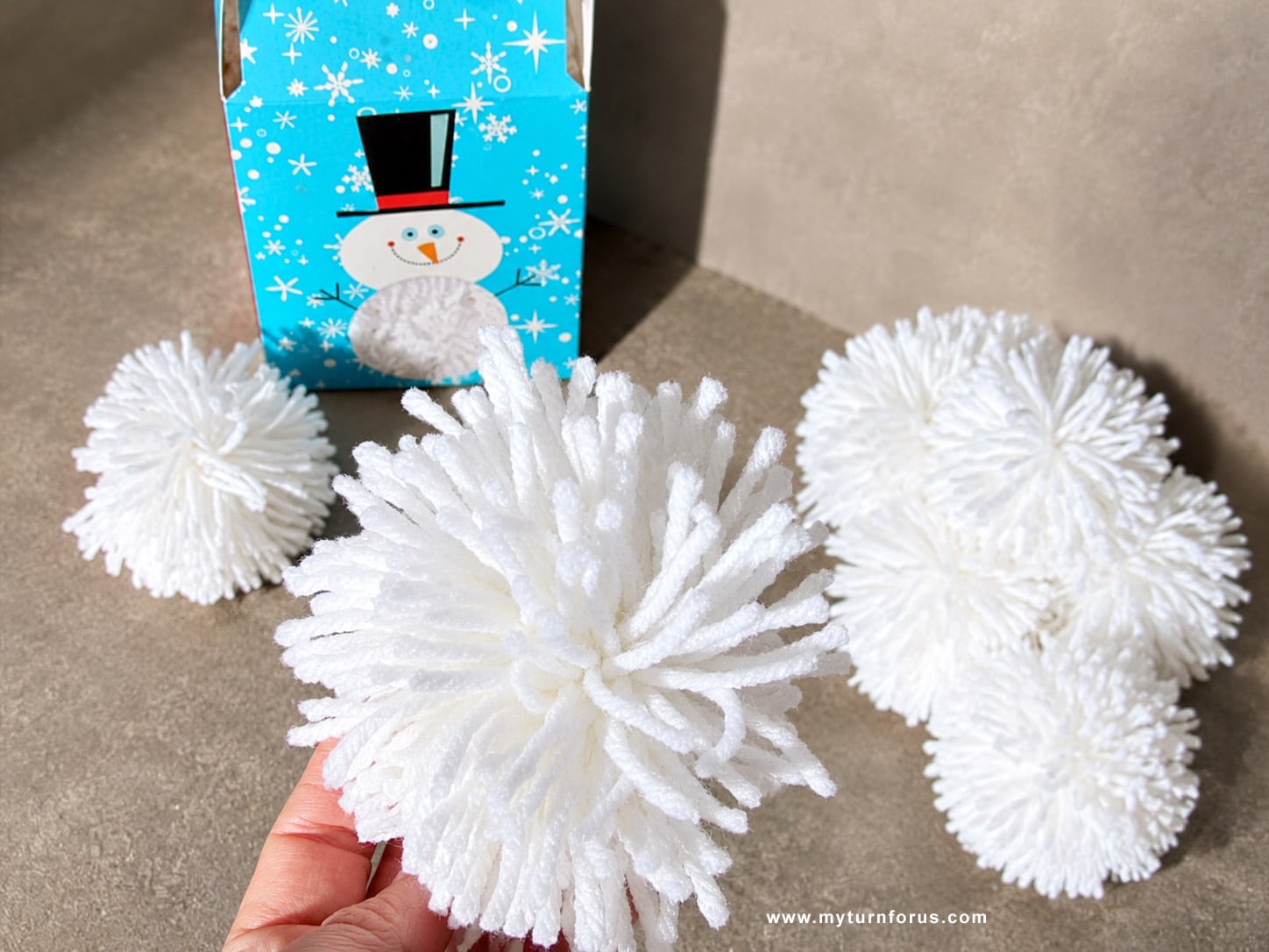 Make these easy yarn pom pom snowballs Close-up of a hand holding a fluffy white yarn pom pom ball with more pom poms in the background.