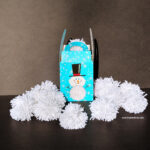 Blue snowman gift box surrounded by white yarn pom pom balls for an indoor snowball fight.