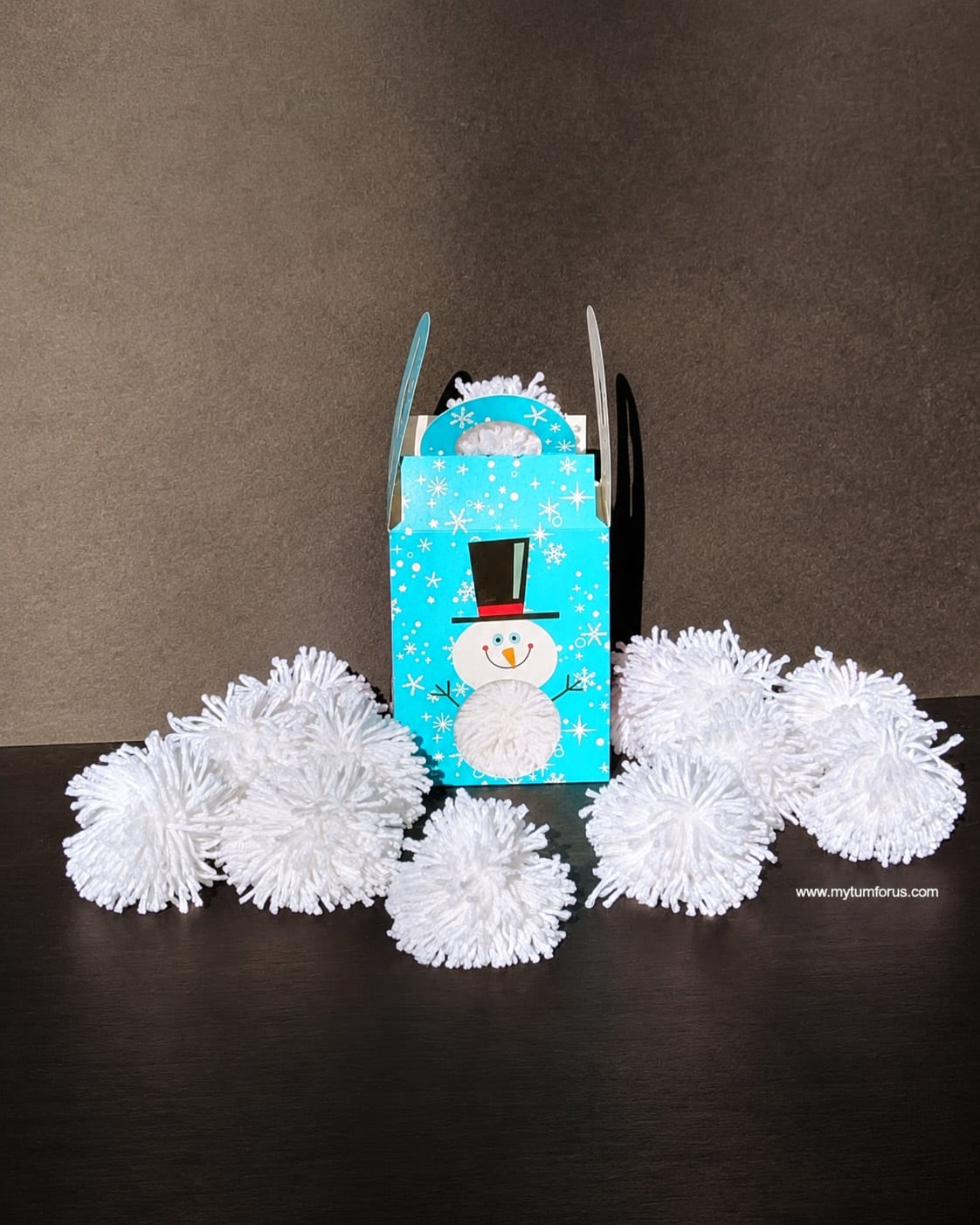 Blue snowman gift box surrounded by white yarn pom pom balls for an indoor snowball fight.
