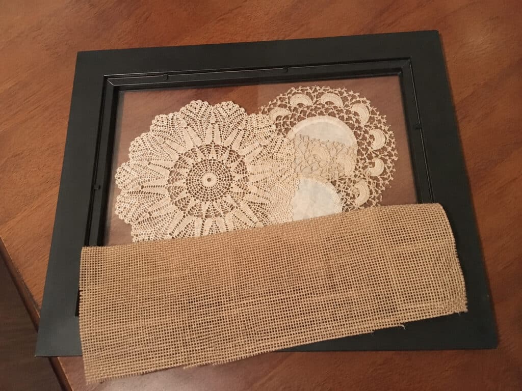 Vintage crochet doilies arranged inside a black picture frame with burlap partially laid over them during assembly.