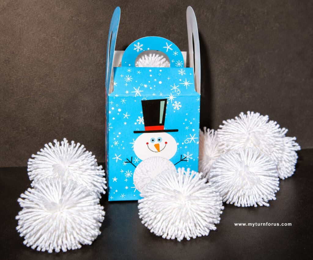 Make this Yarn Pom Pom Indoor Snowball Game White yarn pom pom balls arranged around a snowman gift box on a dark tabletop.